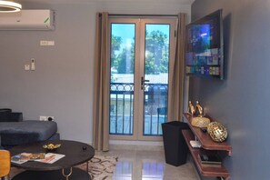 A modern living room with a round coffee table, a sofa, a TV mounted on the wall, and a sliding glass door leading to an outdoor balcony.