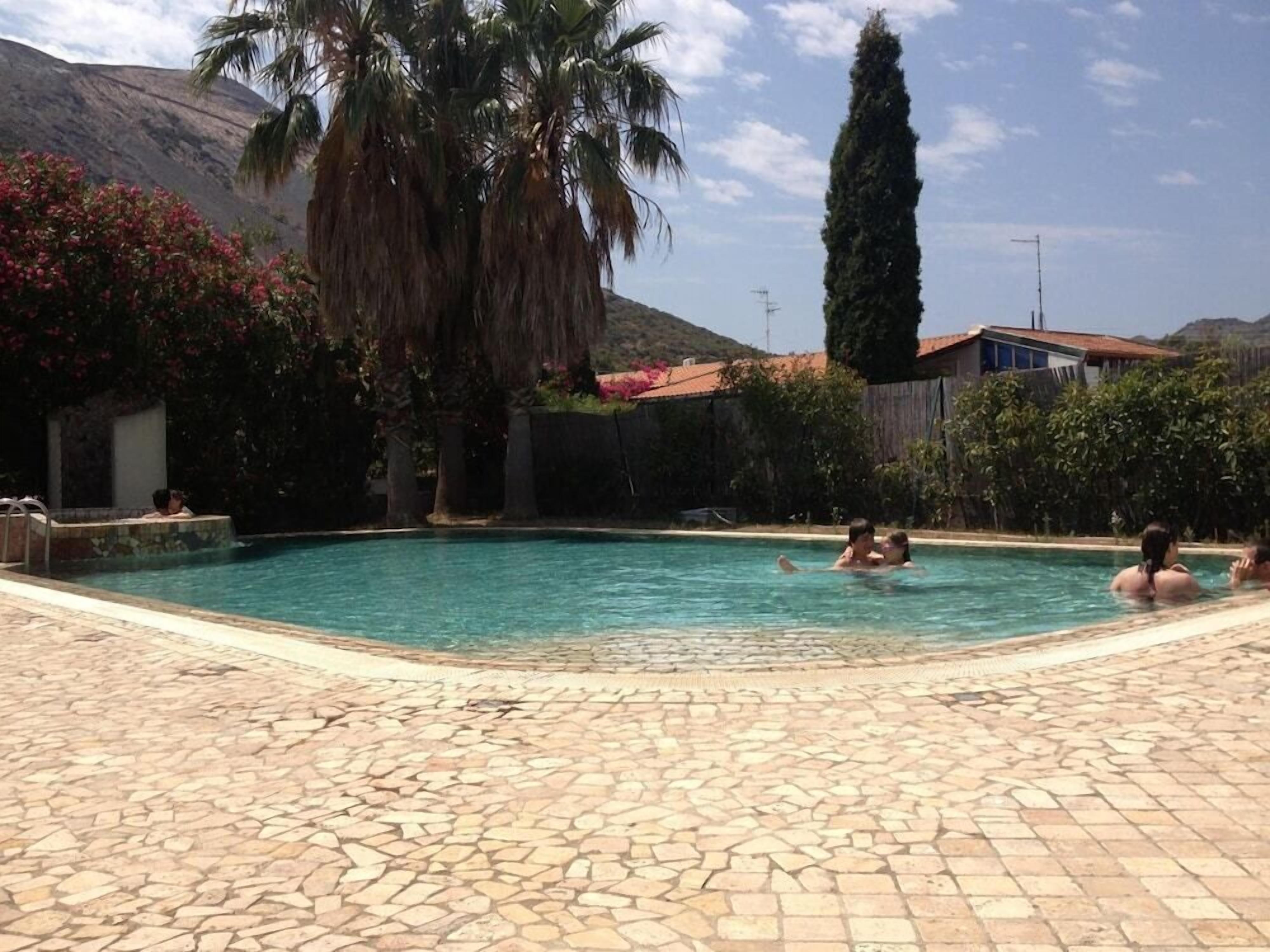 Ein Schwimmbecken mit Steinplatten, umgeben von Palmen und einem Berg im Hintergrund.
