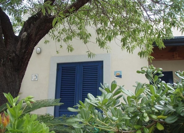 A building with a large tree in front, a blue shuttered window, and a small sign on the wall. - Aura Hotel (Lipari)