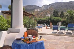 Una mesa con alimentos para el desayuno, asientos al aire libre y un área de piscina con sillones de descanso.