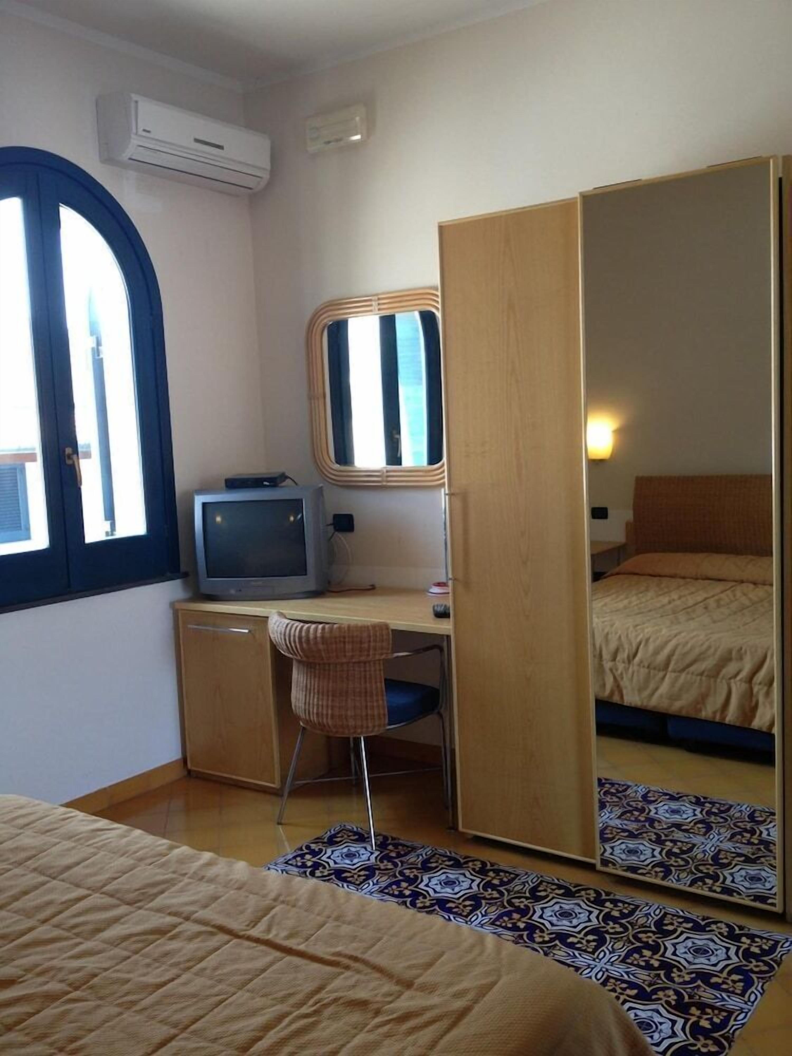 Une chambre à coucher de taille modeste, comprenant un lit, un bureau avec une chaise, une télévision, une armoire et une fenêtre avec des stores.