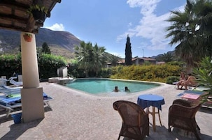 Zona de piscina con sillas de descanso y una mesa, rodeada de vegetación y un paisaje montañoso.