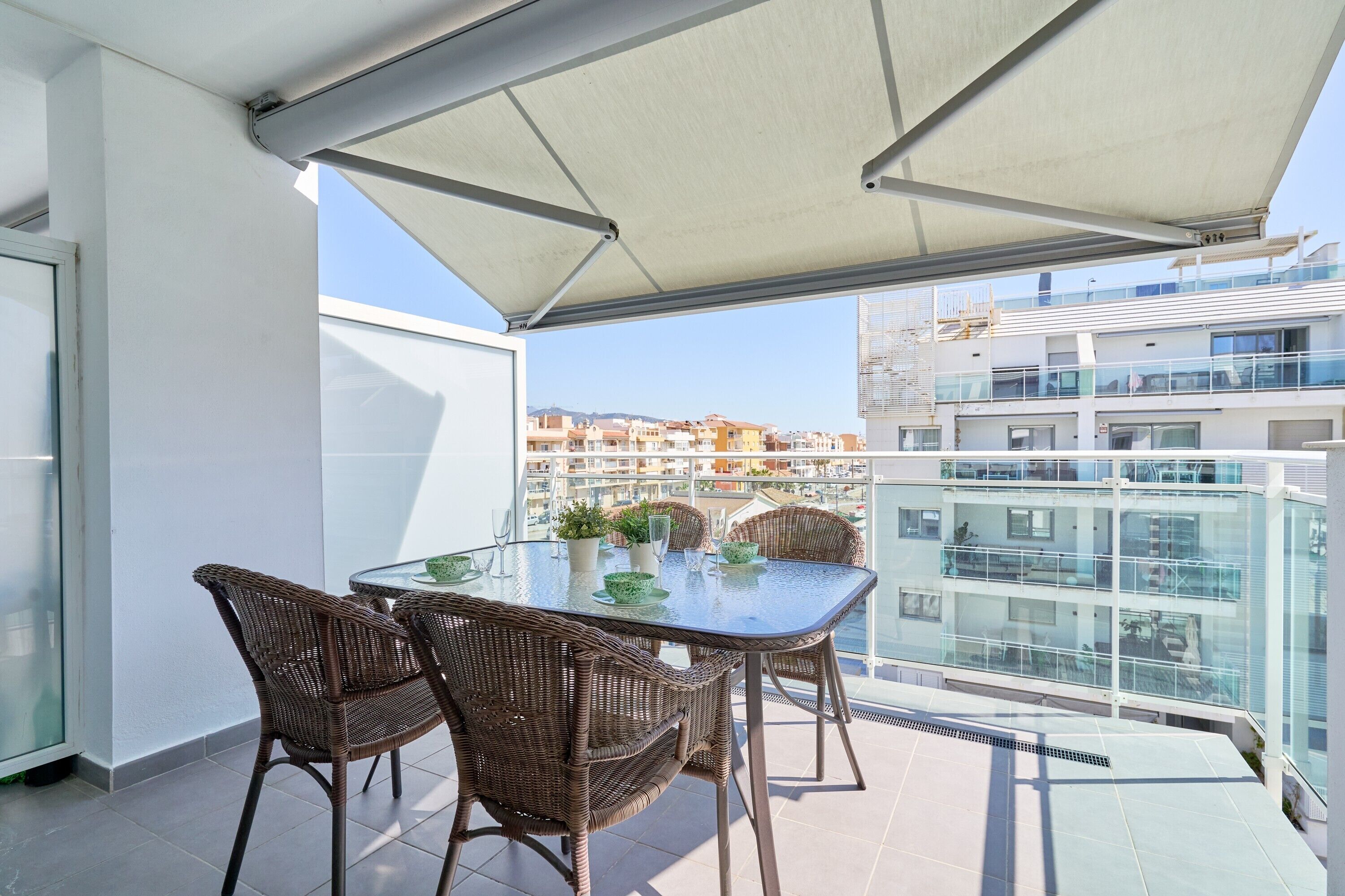 A balcony with a glass table and wicker chairs, overlooking a cityscape with modern buildings.