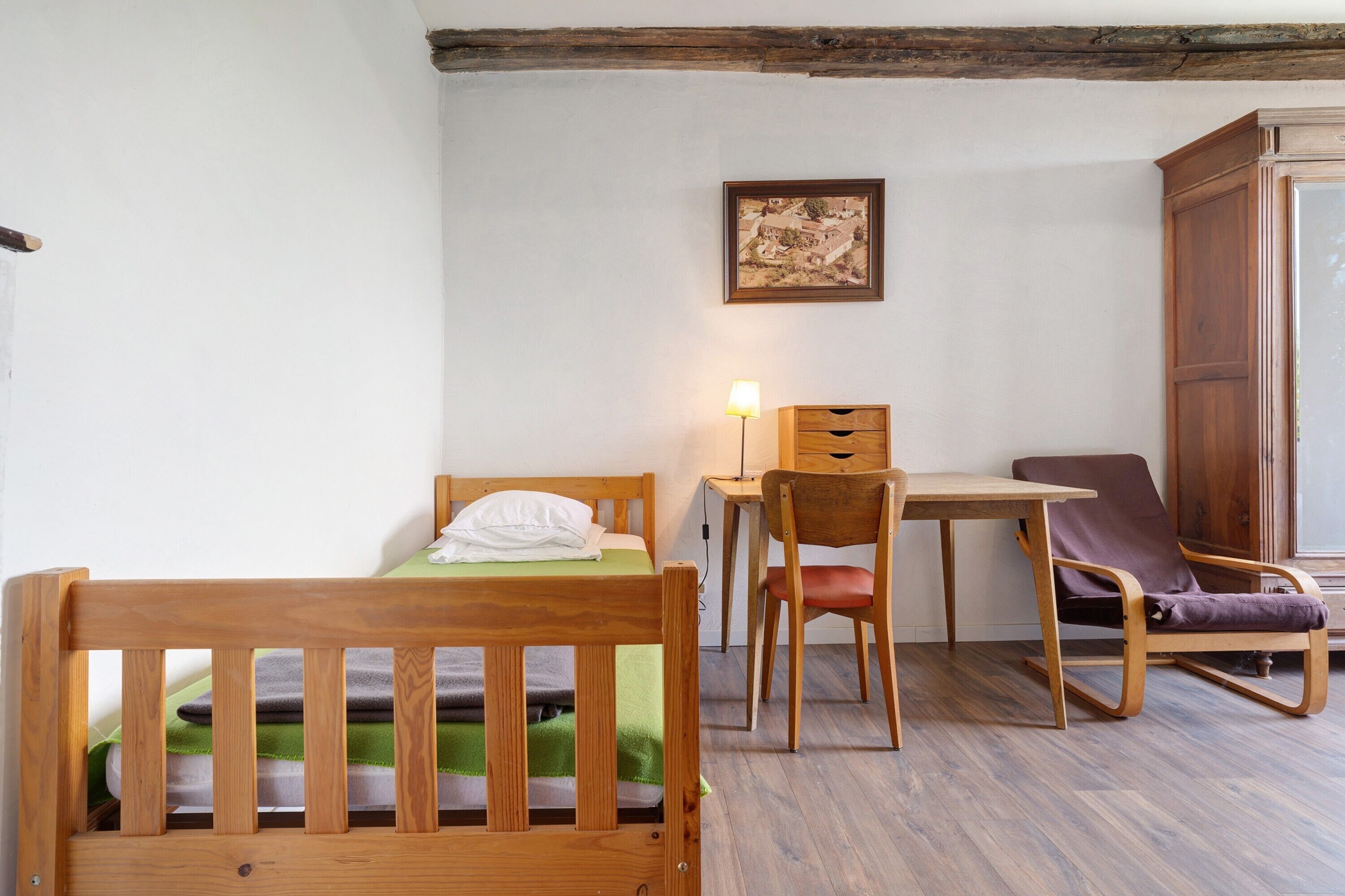 A room with a wooden bed, a desk with a lamp, a chair, and a wardrobe.