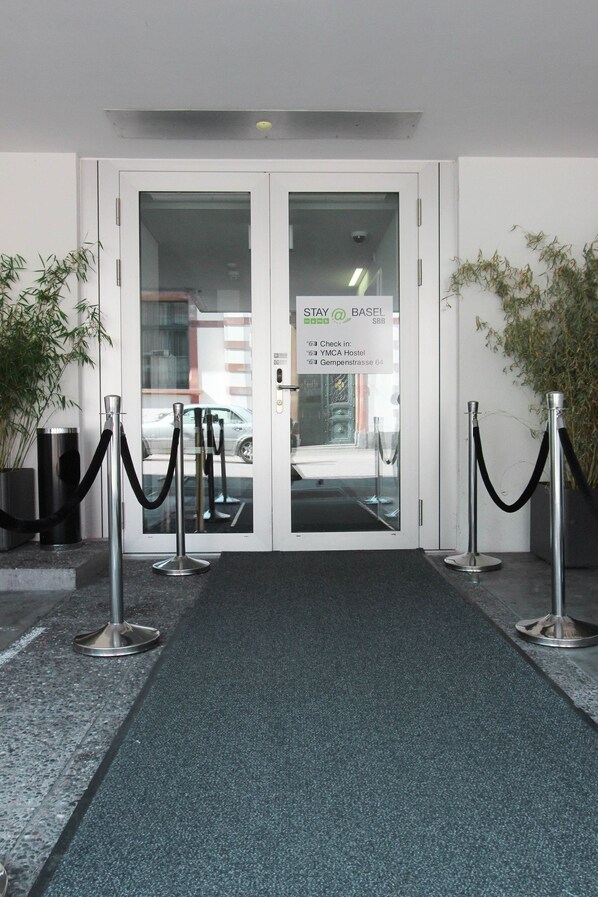 A hotel entrance with a check-in sign, a glass door, a black carpet, and a metal barrier. - Appartements By Stay At (Basel)