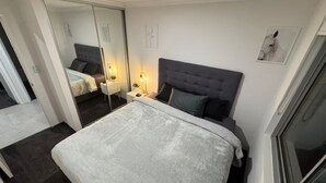 Un dormitorio con una cama grande, una mesita de noche con lámpara, un espejo y un cuadro enmarcado en la pared.