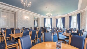Un salón de conferencias de hotel con mesas y sillas redondas, ventanas amplias con cortinas y candelabros.