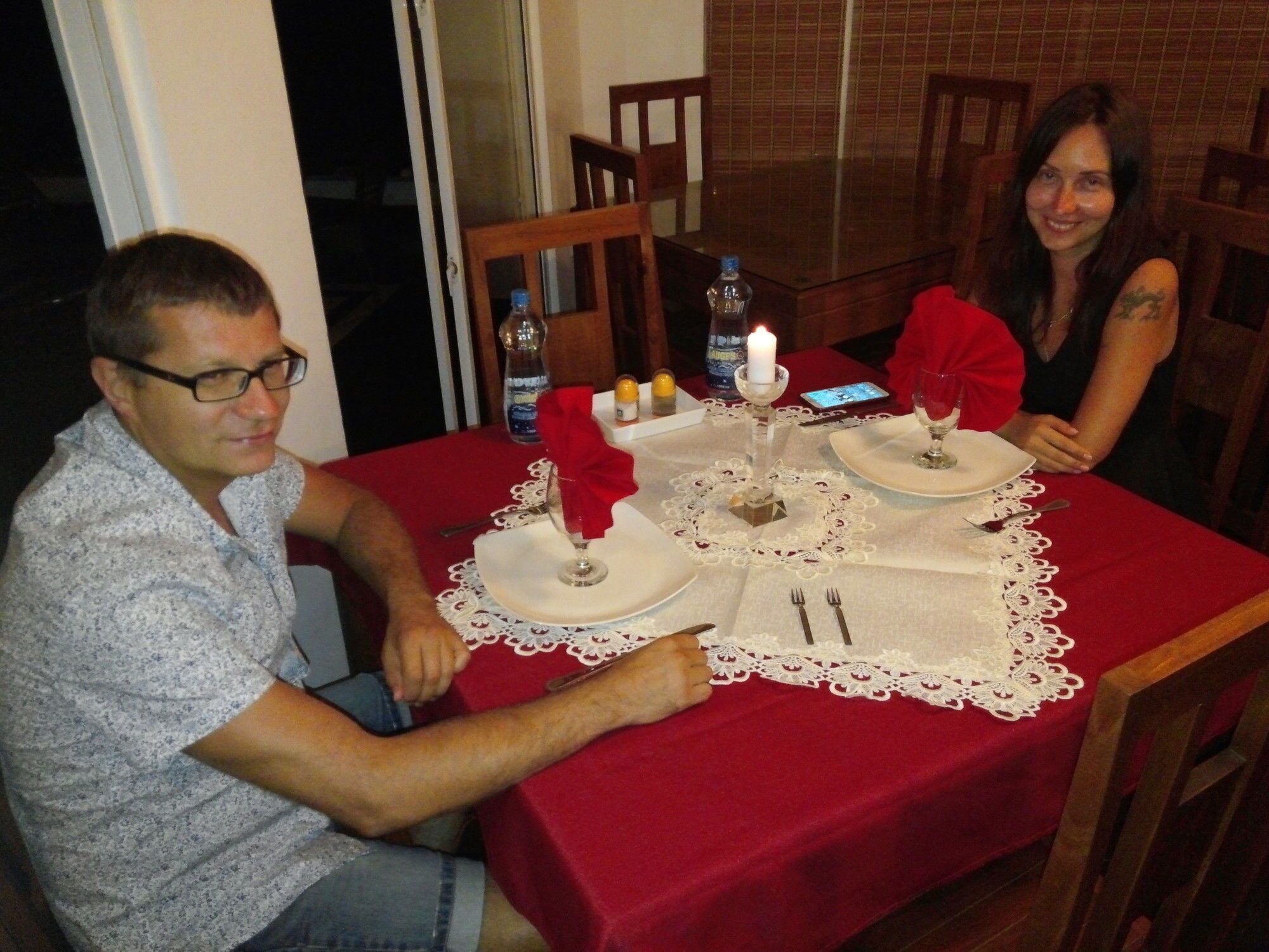 A dining table set for two with a red tablecloth, white lace doilies, and a candle centerpiece.