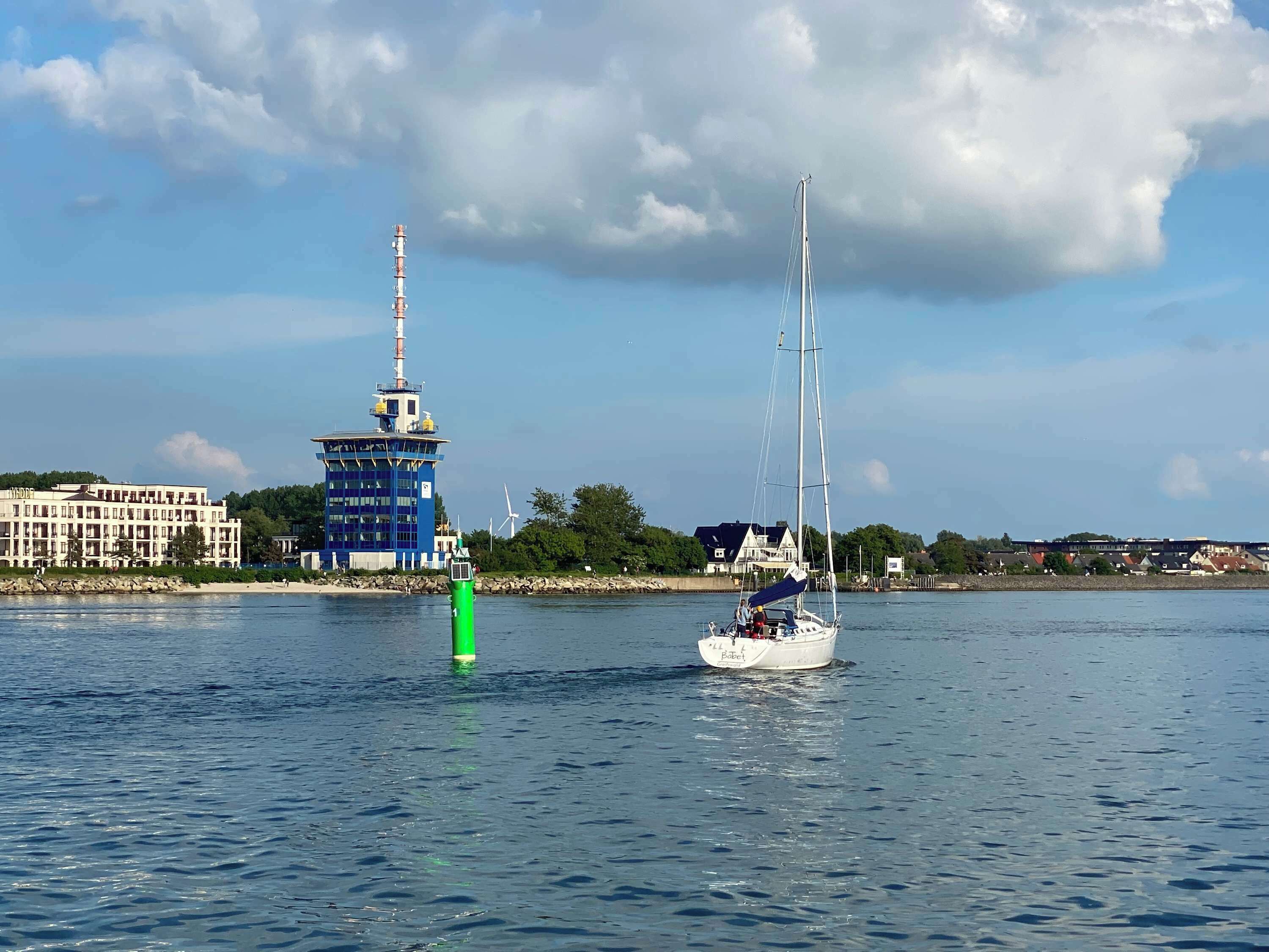 Een kustgezicht met een zeilboot op het water, een vuurtoren en een hotel op de achtergrond.