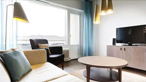 A modern hotel room with a sofa, armchair, and a small round table. There is a television on a cabinet and a window with curtains. - Sato Hotelhome Kamppi (Helsinki)