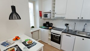 Cocina compacta con gabinetes blancos, microondas, estufa y lavavajillas. Hay una ventana con persianas, una lámpara colgante y una pequeña mesa con un sándwich y naranjas.