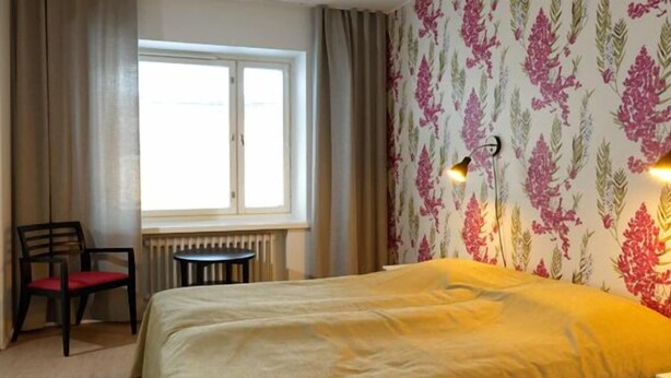 Habitación de hotel con cama, una silla, una mesita, un radiador y una ventana con cortinas.