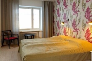 Een hotelkamer met een bed, een stoel, een klein tafeltje, een radiator en een raam met gordijnen.