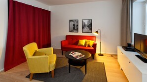 Una sala de estar moderna con un sofá rojo, un sillón amarillo y una mesa de centro. Hay dos fotografías blanco y negro en la pared, una lámpara de pie y un televisor sobre un mueble blanco.