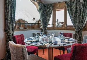 A dining area with a round table set for six, red upholstered chairs, and a view of snow-covered houses through large windows. - Aiguille Percee Hotel Restaurant (Tignes)