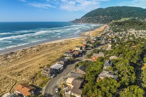 Vue aérienne d’une ville côtière avec des maisons le long de la plage, une route et une colline boisée en arrière-plan.