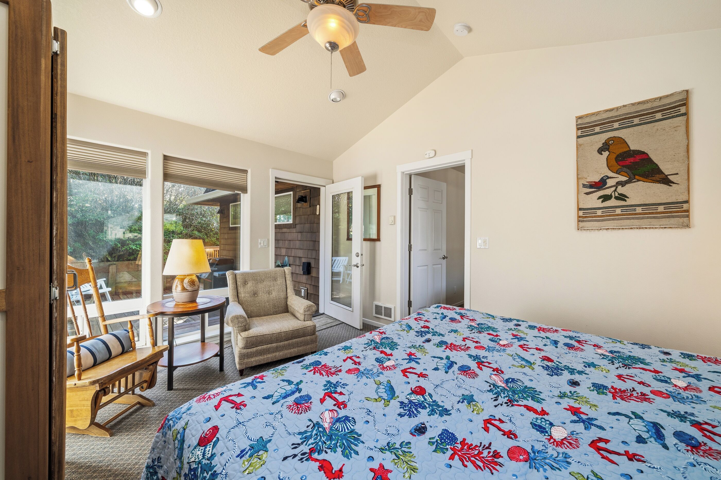 A bedroom with a bed, a ceiling fan, a chair, a table, and a door leading to a balcony.