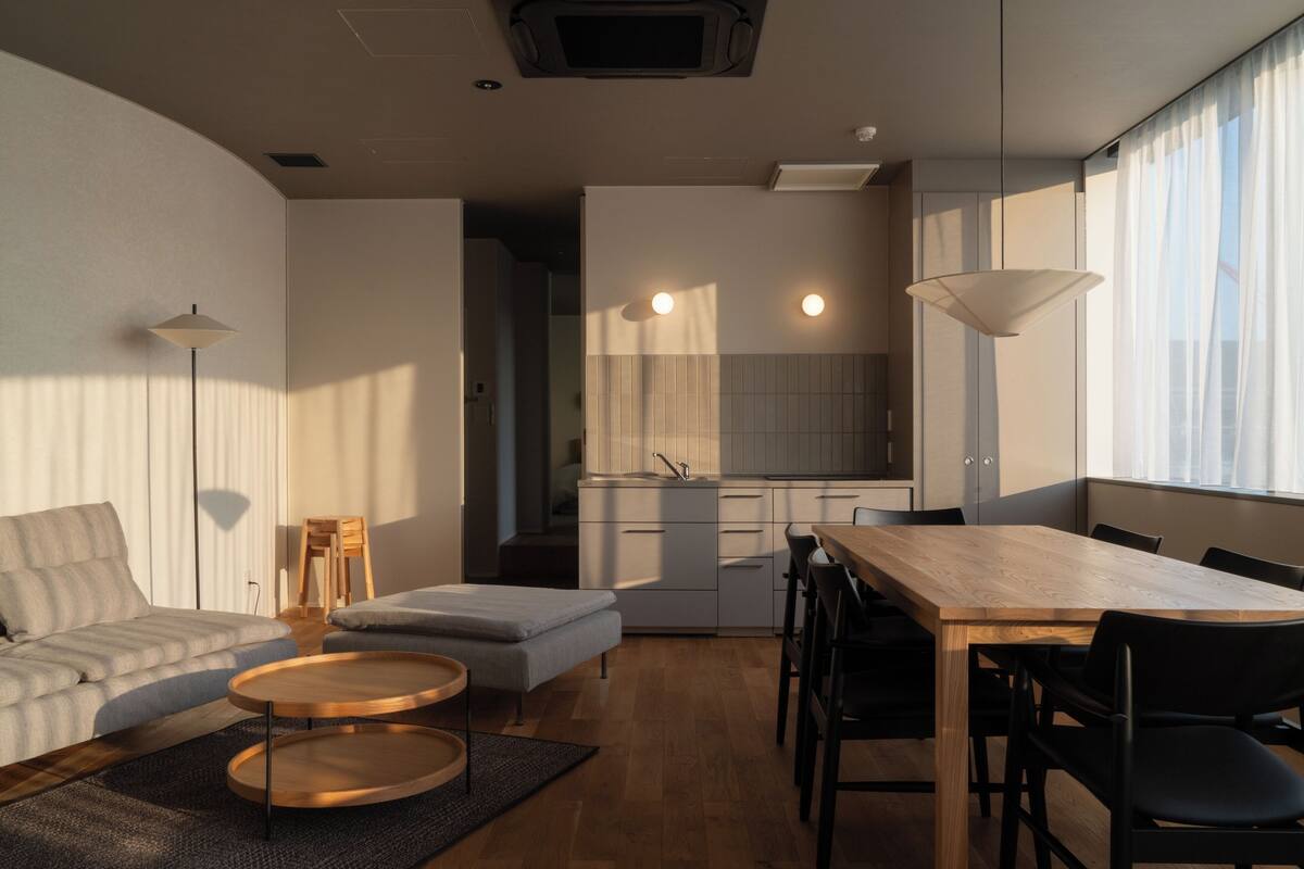 A modern living room with a sofa, ottoman, round coffee table, and a dining table with chairs. There is a kitchen area with a sink and cabinets, and a large window with curtains.