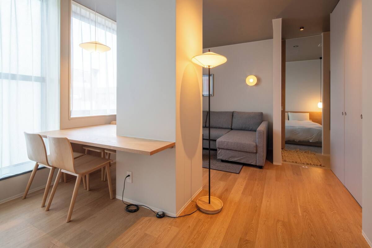 A modern room with a wooden desk, two chairs, a grey sofa, and a bed.