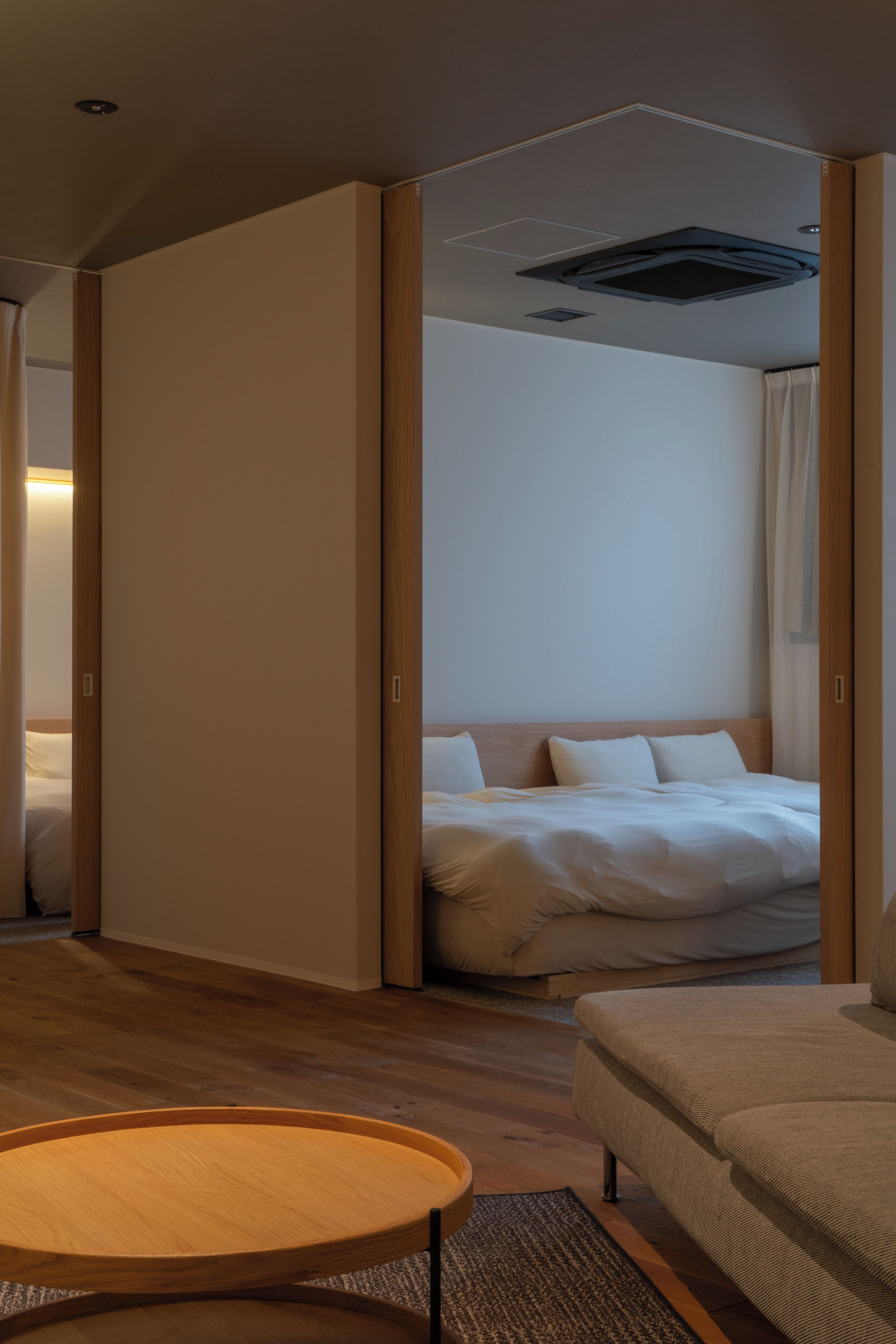A modern hotel room with a wooden floor, a round wooden table, a beige sofa, and a bed with white bedding.