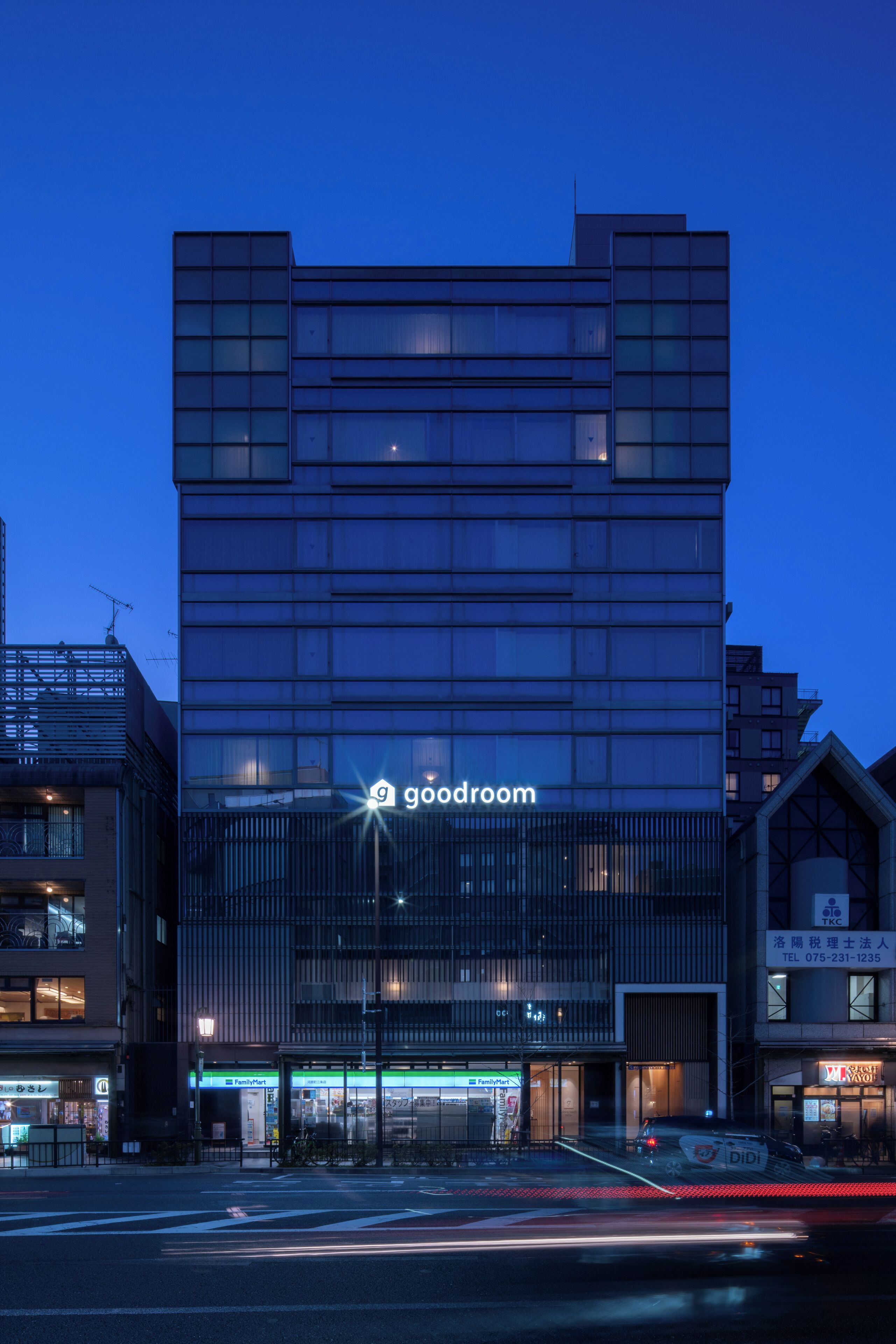 A modern building with a 'goodroom' sign, featuring a glass facade, a ground-level entrance, and a visible street with a crosswalk.