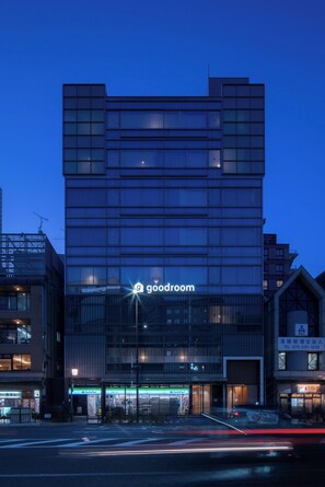 Un edificio moderno con un cartel que dice 'goodroom', con fachada de vidrio, entrada a nivel de calle y una calle visible con un paso de peatones.