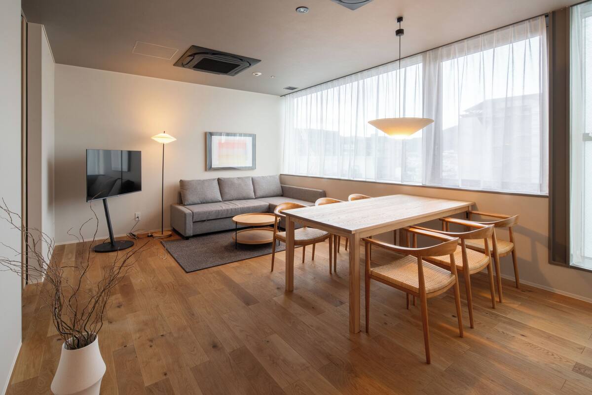 A modern living room with a wooden dining table, chairs, a sofa, a TV, and a large window with sheer curtains.
