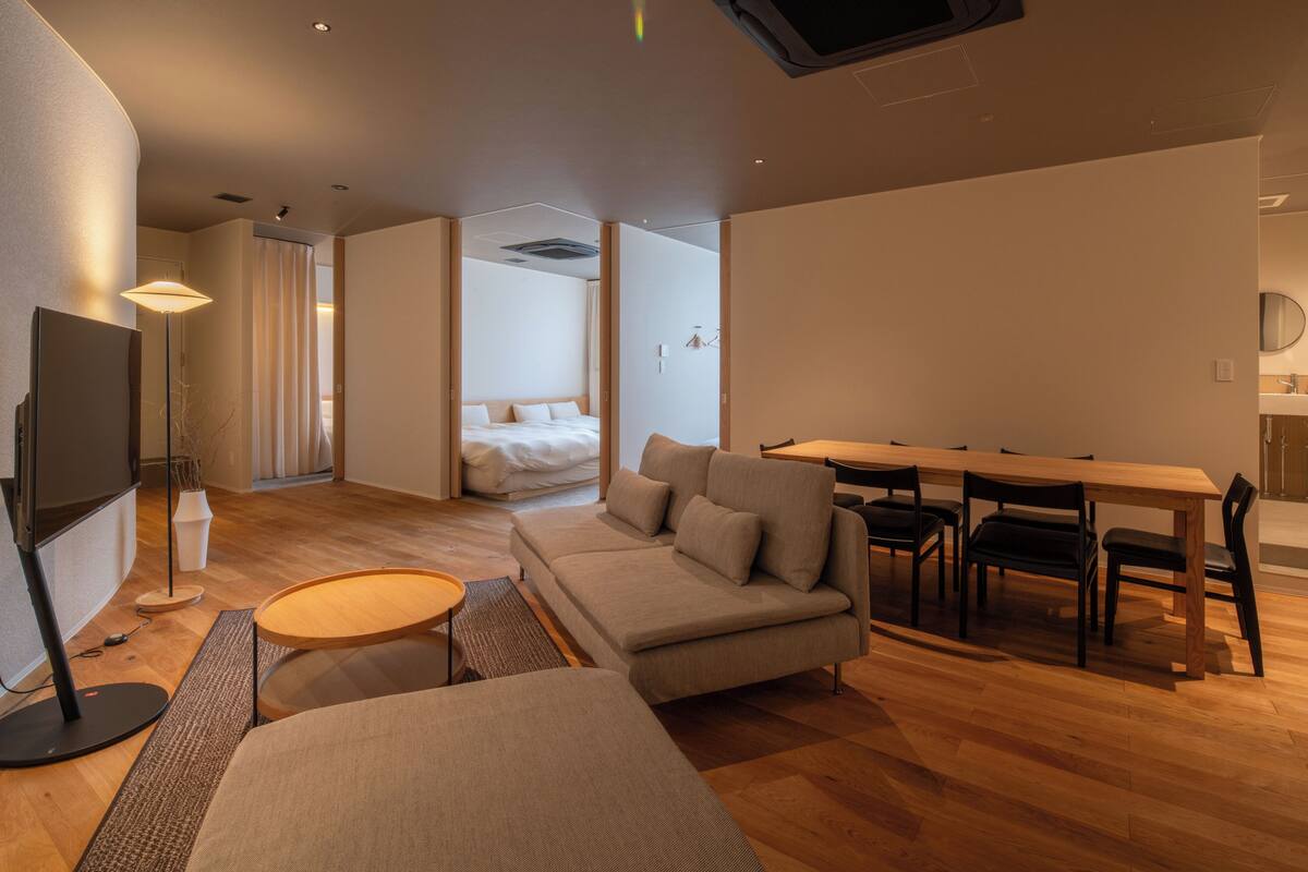 A modern hotel room with a sofa, a dining table with chairs, a TV, and a large window.