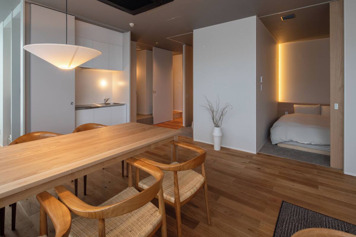 A modern hotel room with a wooden dining table, four chairs, a bed, and a small potted plant.