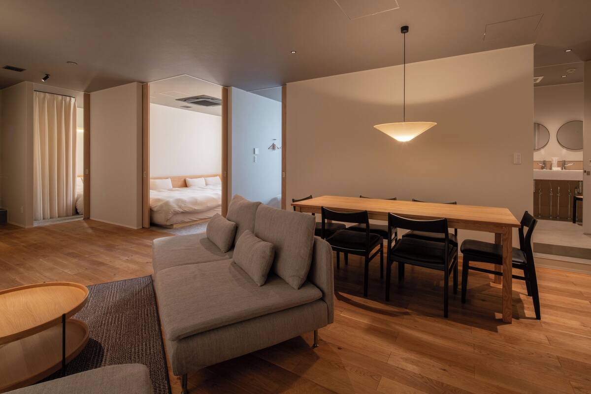 A modern hotel room with a sofa, dining table, chairs, and a bed in the background.