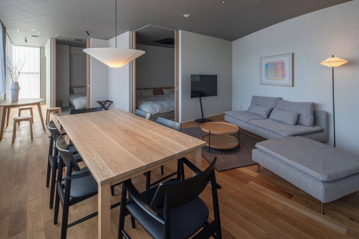 A modern living room with a wooden dining table, grey sofa, and a TV mounted on the wall.