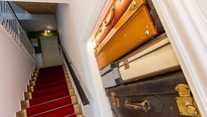 Un escalier recouvert d’une moquette rouge, menant à une pièce où sont entassées des valises.