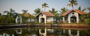 A row of white buildings with red-tiled roofs, surrounded by palm trees and a calm water body.