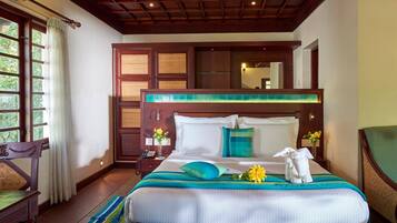 A hotel room with a large bed, a wooden headboard, a nightstand with a phone, a window with curtains, and a view of greenery outside.