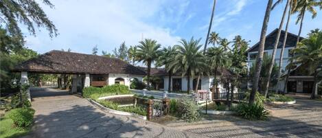 A paved courtyard with palm trees, a covered walkway, and a building with a tiled roof.