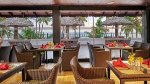 Een restaurant met rieten meubels, tafels met rode servetten en uitzicht op palmbomen en de zee.