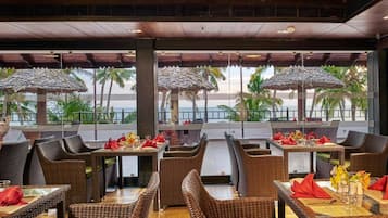 Ein Restaurant mit Korbmöbeln, Tischen mit roten Servietten und Blick auf Palmen und das Meer.