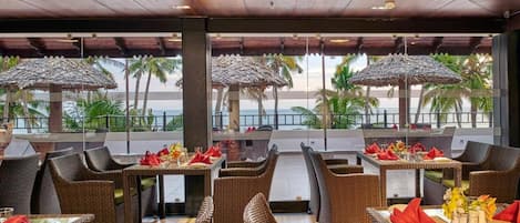 A restaurant with wicker furniture, tables with red napkins, and a view of palm trees and the sea.