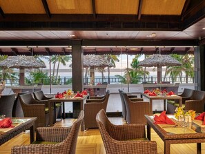 A restaurant with wicker furniture, tables with red napkins, and a view of palm trees and the sea.