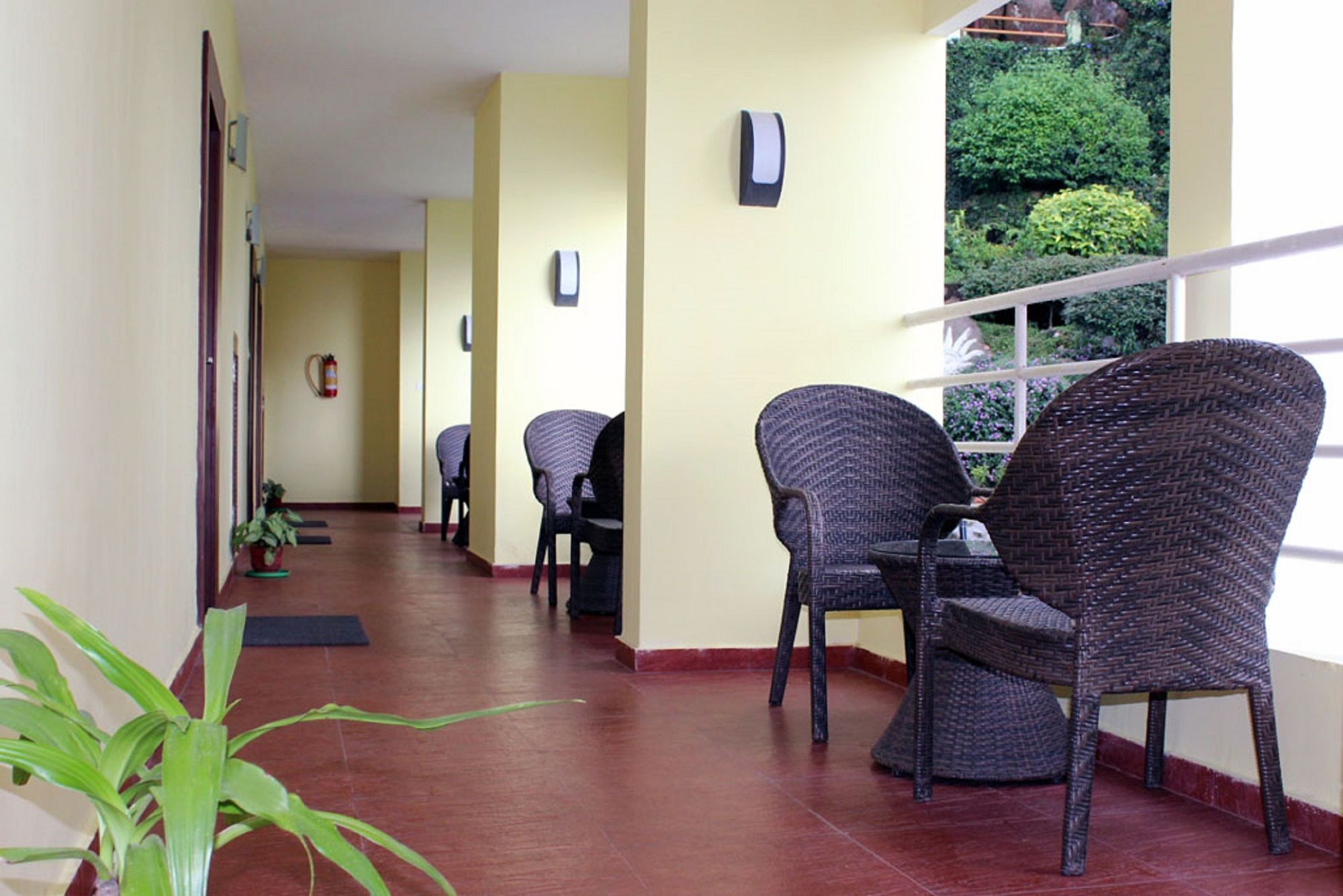 A corridor with wicker chairs and a table, a fire extinguisher, and a window with a view of greenery.