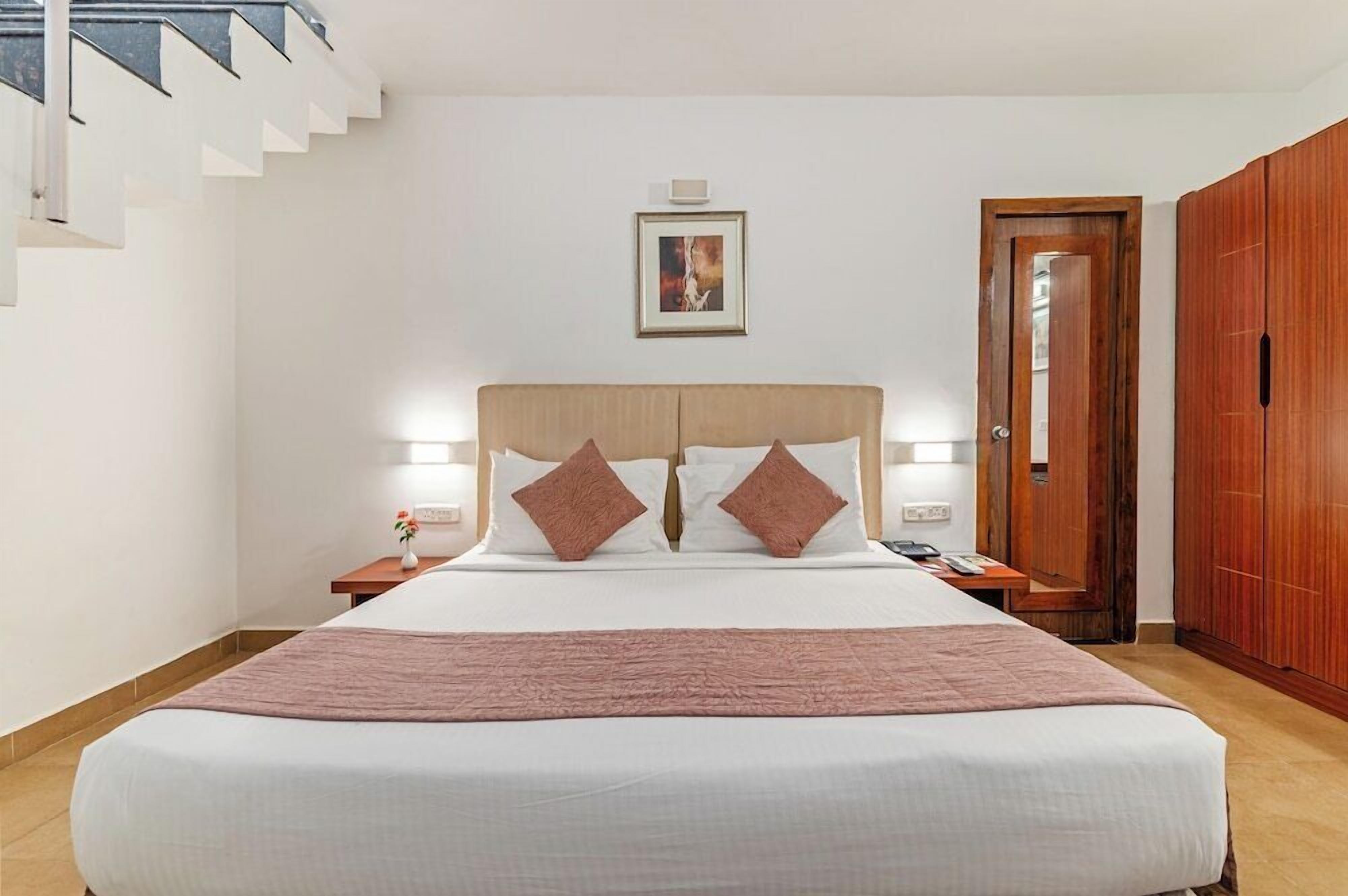 A hotel room with a large bed, a wooden headboard, a nightstand with a lamp, a framed picture on the wall, and a wardrobe.