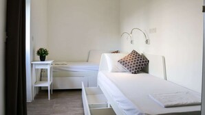 A small, clean room with two beds, a bedside table with a plant, and a wall-mounted light fixture. - Inhawi Hostel (St. Julian's)