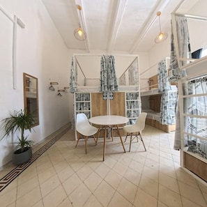 Une chambre avec un lit superposé, une table ronde avec des chaises, une plante en pot et une étagère agrémentée de rideaux à motifs.
