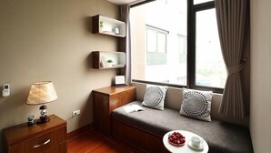 A cozy room with a wooden nightstand, a lamp, a small table with a bowl of fruit and a cup, a sofa with patterned cushions, and a window with curtains. - Istay Hotel Apartment 1 (Hanoi)