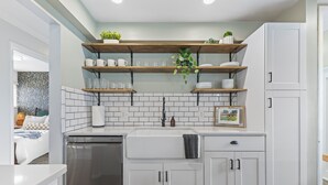 A modern kitchen with white cabinetry, a stainless steel dishwasher, and wooden shelves with plants. - Luxury Look in Plymouth I Walk to Downtown l Sleeps 8 I Hot Tub (Plymouth)