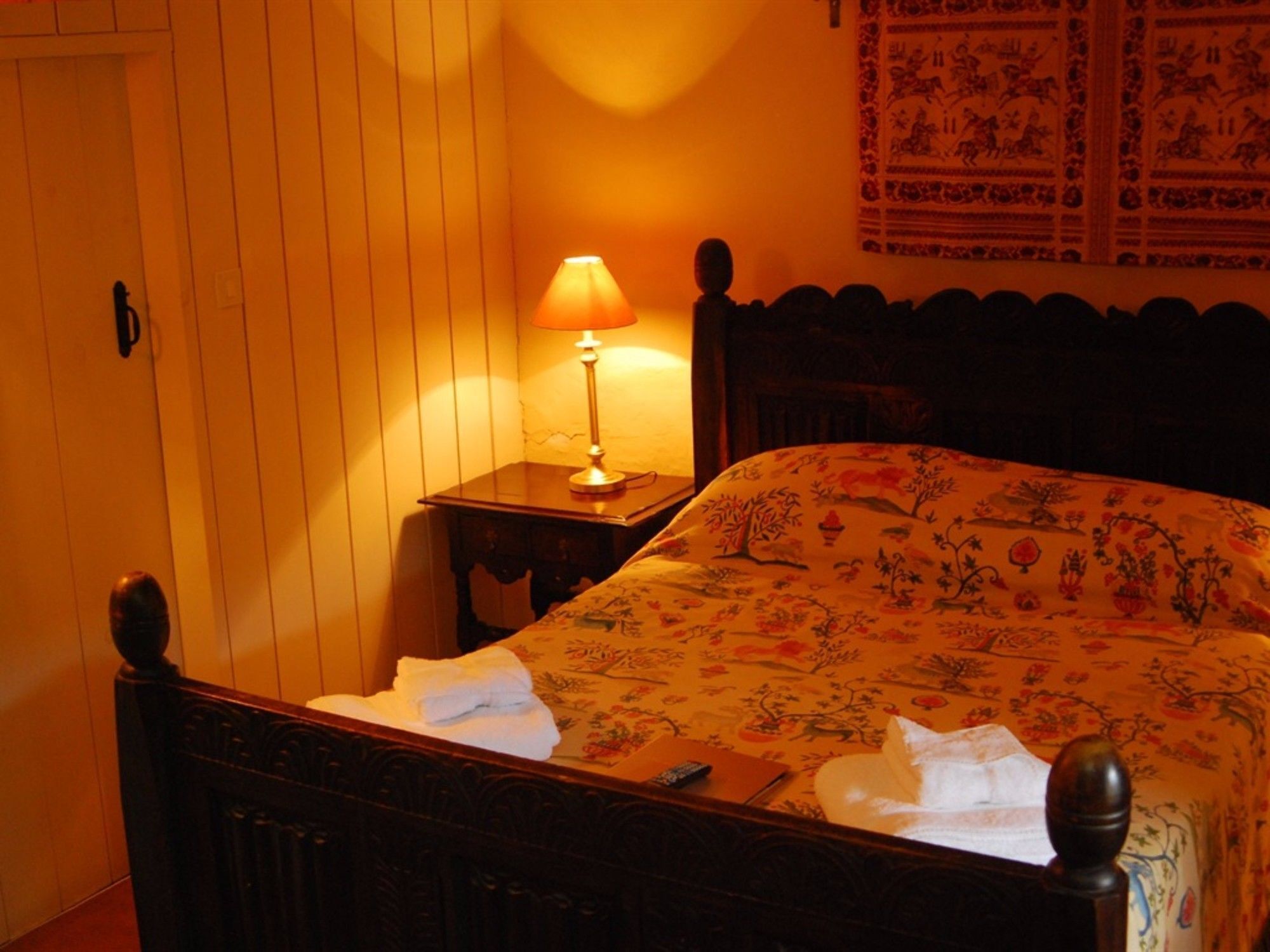 A bedroom with a wooden bed, a nightstand with a lamp, and a wall with a tapestry.