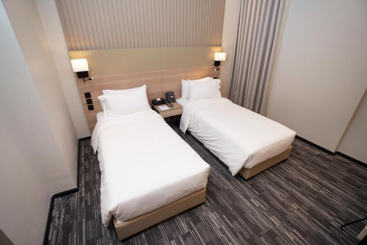 Habitación de hotel con dos camas individuales, una mesita de noche con teléfono y cabecero con líneas verticales.