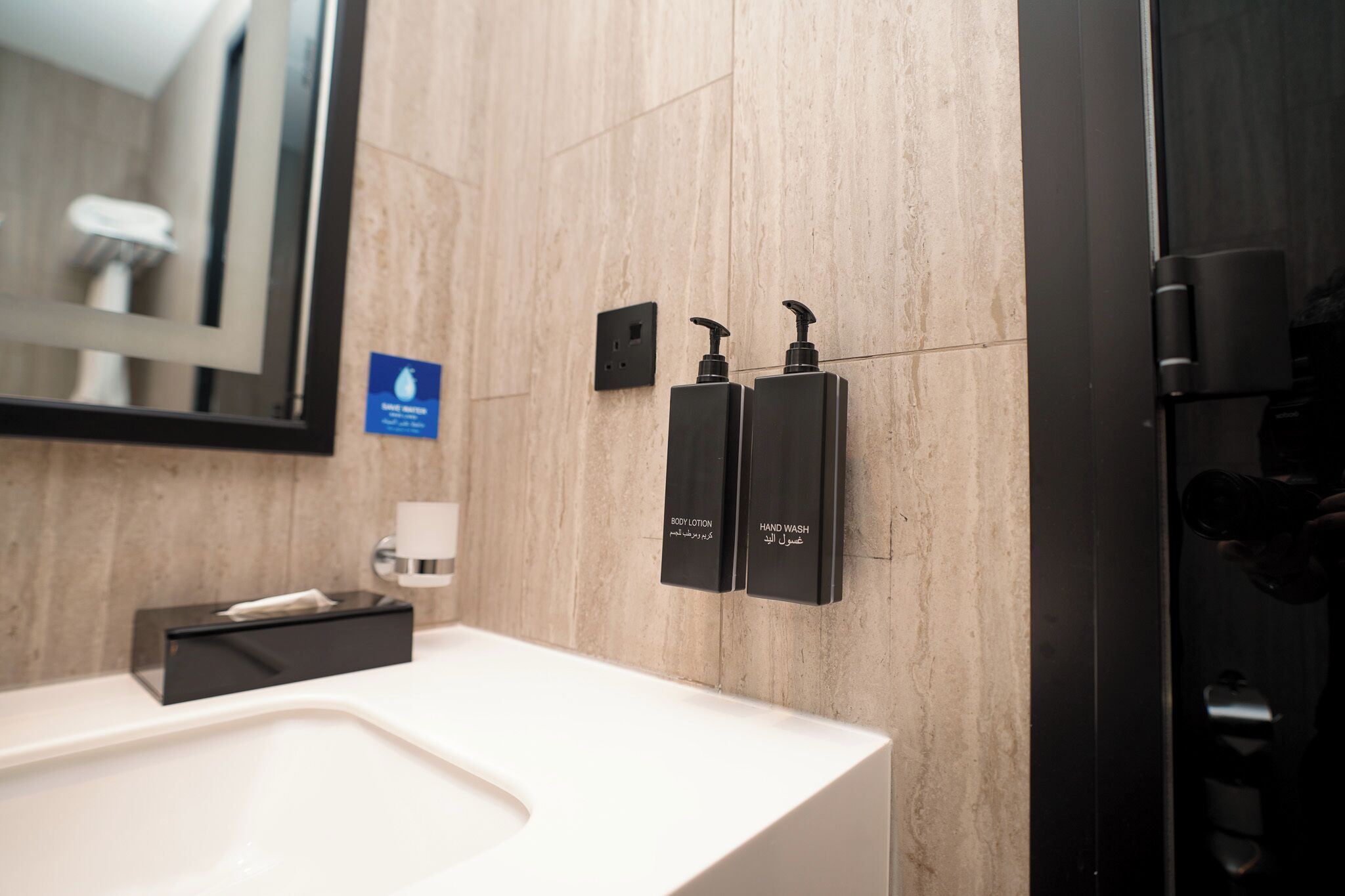 A modern bathroom with a white sink, a mirror, and two black dispensers labeled for body lotion and hand wash.