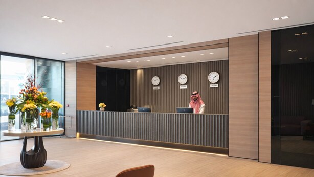 Uma área de recepção moderna com um recepcionista, um relógio exibindo a hora em três fusos horários e uma mesa com arranjos florais.