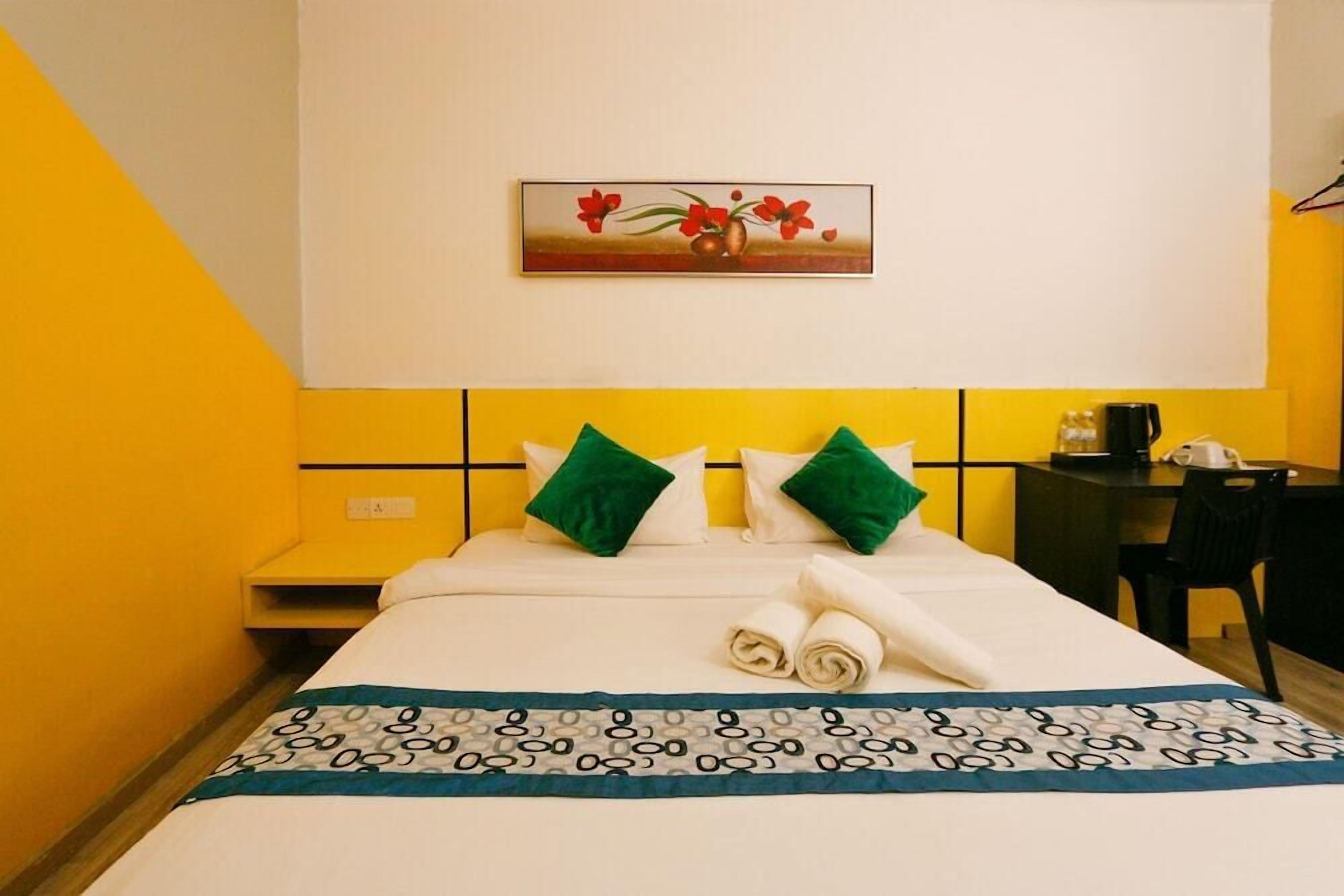 A hotel room with a bed, green pillows, a painting of flowers, and a desk with a chair.